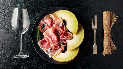 Prosciutto, melon, and rosemary on a black plate.