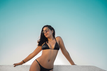 Joven modelo femenina con lentes de sol morocha bronceada en bikini negro posando sobre una baranda...