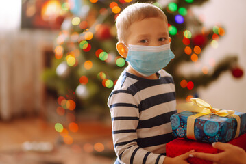 Happy child in Sterile medical mask holding Christmas gifts. Funny little boy posing with presents in Christmas interior design. Winter holidays  in covid-19 pandemic.