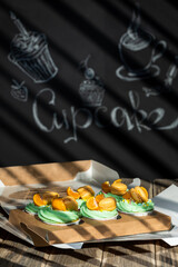 Cupcakes with fruits and berries in a box on a wooden table, a wall with chalk drawings, different light effects.