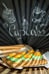 Cupcakes with fruits and berries in a box on a wooden table, a wall with chalk drawings, different light effects.