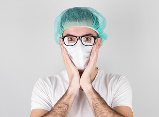 Doctor surgeon in white medical mask and a medical cap stands over white background with surprise,holding face with the hands. Сoncept of covid 19, flu and seasonal cold.