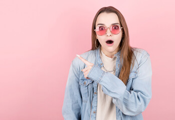 Horizontal picture of young female isolated on pink background wearing red eyeglasses, pointing leftwards with forefinger with surprise.