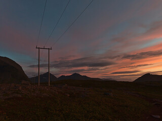Wooden power pylons in rocky landscape after sunset with dramatic orange and pink colored sky and mountains in background on Kvaløya island in northern Norway in late summer.
