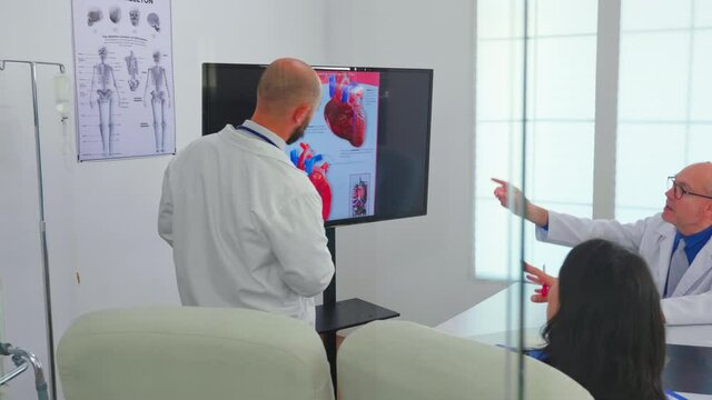 Experienced man doctor discussing about heart issues using digital image, together with cvalified colleagues in meeting room, pointing on monitor. Doctors analysing diagnosis for treatment of patients