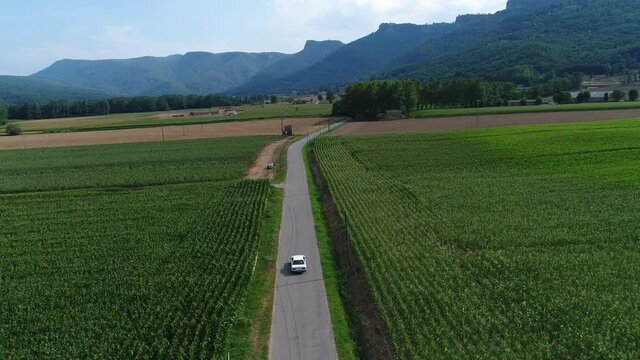 White Classic BMW Car Travelling Close To Hostalets de Bas In Olot Catalonia, Spain Passing By Wide Green Fields Background With Foggy Mountains Under The Cloudy Sky - Aerial Shot