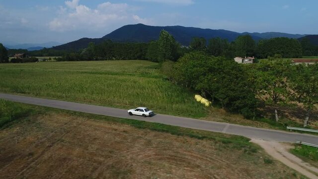 White BMW Car Driving Along Green Cornfields In Hostalets De Bas In Catalonia, Spain. - aerial pullback