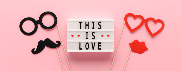 White light box with text This is Love and paper props mustache, lips and glasses on pastel pink background. Concept Valentine's Day. Creative flat lay, banner