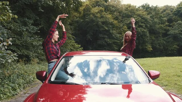 Girfriends enjoying time together. Two female friends sitting from ides of the red car haing hand up in the air while riding in the car. Weekend activities.