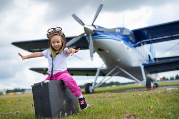 A little girl in a pilot's costume carries a retro suitcase and walks along the airfield. A child in a hat and glasses is going on a trip by plane.