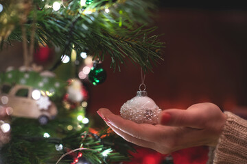 Woman decorating branches of christmas tree with toy at home near fireplace. Cropped, close up