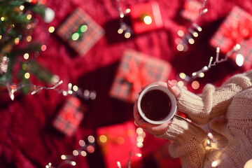 Woman with cup of hot chocolate and wrapping christmas gifts boxes for family on fluffy plaid near christmas tree. Top view through the glowing garland.