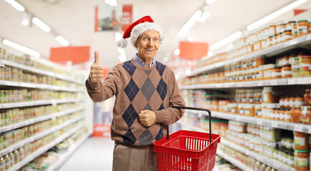 Elderly man with a shopping basket in a supermarket showing a thumb up sign amd wearing a santa...