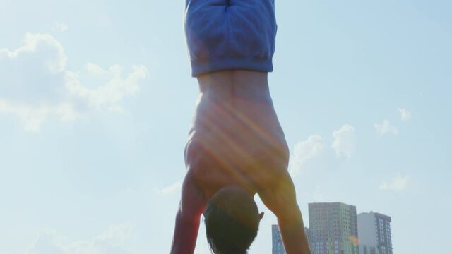 A man does a street workout at a sports ground. A sportsman does a handstand on parallel bars against an urban landscape