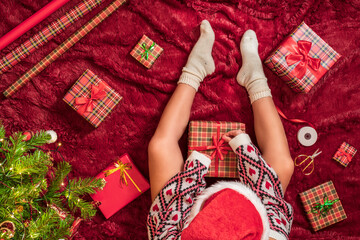 Top view of woman in red santa claus hat wrapping christmas gifts boxes for family sitting on fluffy plaid near christmas tree. Flat lay.