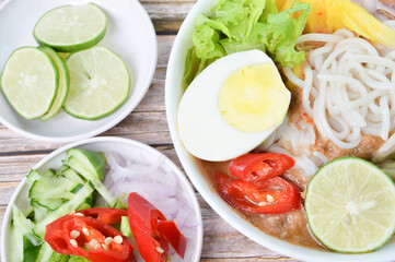 Close up view of Noddle in Tangy Fish Gravy or Laksa Utara is a Special Malaysian Food Popular in Penang in a white bowl served with chopsticks, egg, lime, cucumber, chilies, pineapples and salads.