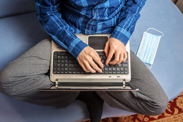 A man is typing on a laptop while sitting on the couch. The medical mask is nearby.