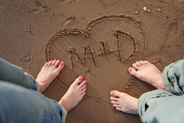 couple standing by heart with initials scratched into beach showing feet and legs only