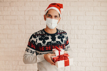 Happy man in medical face mask, Christmas sweater hold gift boxes with red bow in hands. Young festive man in Santa hat, face mask with New Year presents. Celebrate winter holidays. Merry Xmas Eve