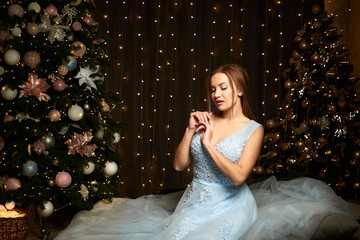 A woman in an evening blue dress sits near a Christmas tree and dreams. Lots of lights and...