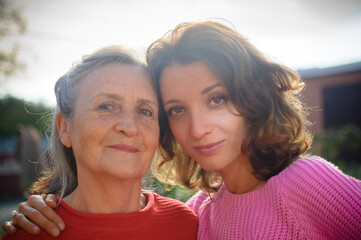 Obraz premium Senior mother with gray hair with her adult daughter looking at the camera in the garden and hugging each other during sunny day outdoors