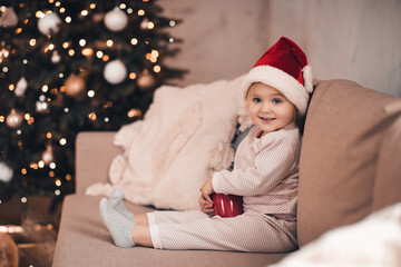 Obraz na płótnie Canvas Smiling baby 1-2 year old wearing pajamas and red santa claus hat sitting over Christmas tree over glowing lights close up. Winter holiday season. Celebration.