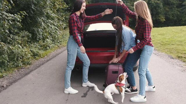 Three teenage girls are going on a trip. One girl is loading the suitcase into a car boot. Friends travelling,time together.