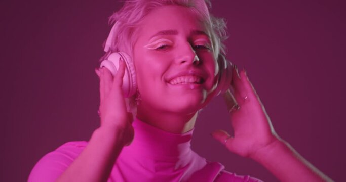 Portrait of active young woman with unusual appearance, listening music in headphones, dancing, feeling excited, moving body energetically. Pink neon light background.