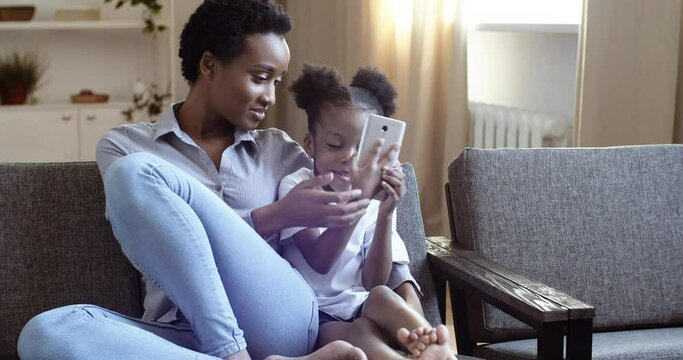 Young mom with short afro hairstyle hugs little daughter curious mixed race child black girl woman shows teaches how to use mobile smartphone gadgets, family takes video making photo sitting on sofa