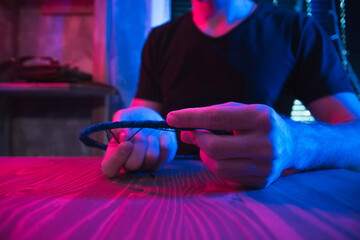 Craftsman makes dreamcatcher in dark workshop illuminated by red and blue neon lighting. Artisan in art studio. Night activity and creativity concept, close up.