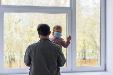 Father and infant daughter wearing protective face masks in a hallway at the hospital clinic during coronavirus pandemic. Patients waiting for a doctor's 
pediatrician consultation  at a large window