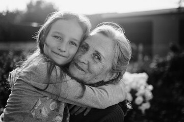 Black and white portrait of senior grandmother with gray hair with her little granddaughter hugging in the garden and during sunny day outdoors, mother's day