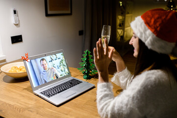 Video call at Christmas eve. A young woman in santa hat greeting a guy on the computer screen. A girl with a glass of champagne sitting at cozy home using laptop and talking online in the evening