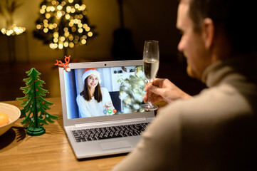 View above shoulder a guy with a glass of champagne is talking online with a cheerful woman in santa hat on the laptop screen. Video call, virtual celebration of Christmass eve