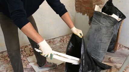 a man in black clothes and cotton gloves collects construction waste in large plastic bags in a...