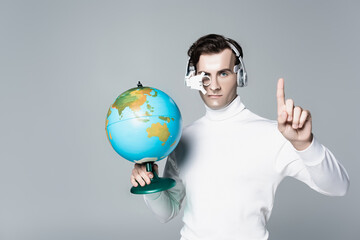Cyborg in headphones holding globe while pointing up with finger isolated on grey