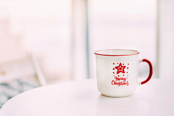 Soft focus white cup with red text and a Christmas star stands on the table by the window. Blurred background. Horizontal photo. Place for the text.