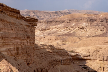 Fototapeta premium Desert landscape with sandy hills and high cliff above dry riverbed Hatira, Negev desert, Israel. Travelers standing on the cliff viewing the panorama of picturesque mountain landscape.