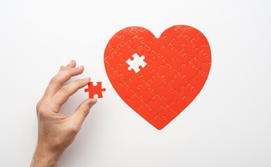 Top view of hand holding last piece near unfinished puzzle in form of heart on white background