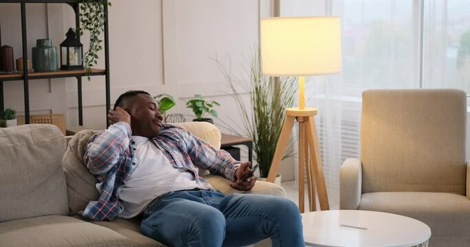 Bored or restless african american man checking his mobile phone and going to sleep on sofa at home