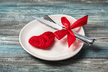 A fork with a knife tied with a red bow and a heart in a plate on a wooden background. The concept of Valentine's day. Side view.