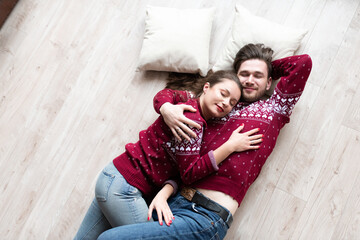 Husband and wife in identical knitted sweaters have fun together celebrating Christmas. The couple hugs and lies on the floor.