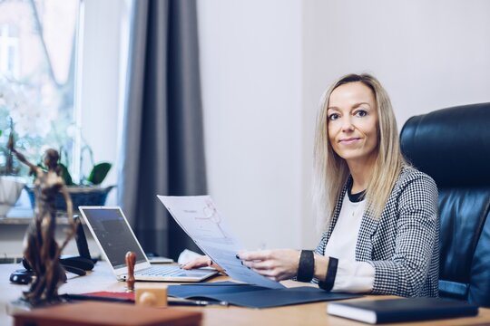 Smiling Female Notary Sitting At The Wooden Desk In The Notary Public Office And And Reading Documents.