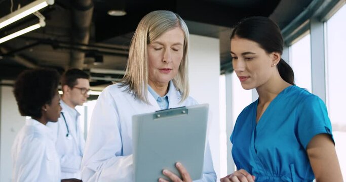 Close up portrait of senior Caucasian female doctor talking to young pretty nurse assistant intern and writing down treatment for a patient. Healthcare workers working in modern clinic