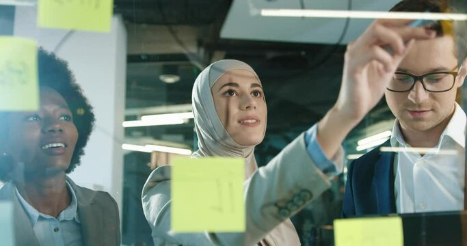 Close up of multi-ethnic male and female professional managers working in modern office. Beautiful Arabian female writing on glass board and talking to team while man typing on tablet Business concept