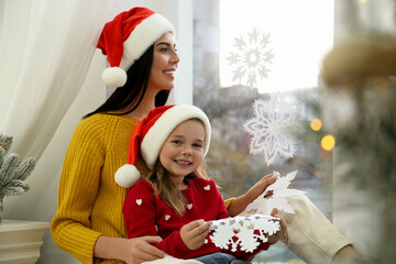 Mother and daughter in Santa hats with paper snowflakes near window at home