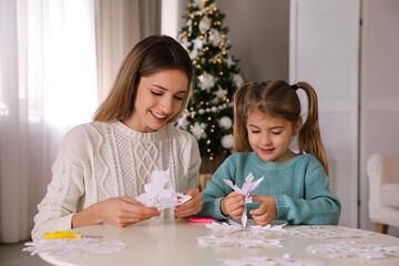 Happy mother and daughter making paper snowflakes at table near Christmas tree indoors