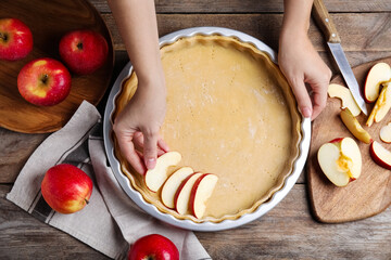 Woman putting apple slice into baking dish with dough to make traditional English pie at wooden...