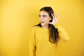 Pretty girl wearing a yellow sweater standing over isolated yellow background surprised with hand over ear listening an hearing to rumor or gossip