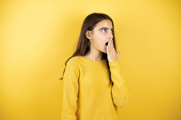 Pretty girl wearing a yellow sweater standing over isolated yellow background surprised with her hands over her mouth